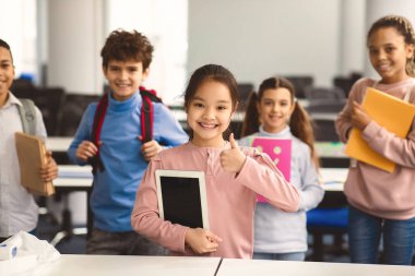 Eğitim ve Teknoloji Konsepti. Elinde dijital tablet olan gülümseyen küçük Asyalı kız ve işaret işareti gösteriyor. Arka planda sırt çantaları olan çeşitli uluslararası sınıf arkadaşları