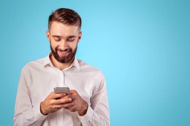 Akıllı telefon ekranına bakan neşeli genç adam, internete göz atıyor, online oyun oynuyor, mavi stüdyo arka planında alışveriş yapıyor. Modern mobil cihaz kullanan mutlu beyaz adam.