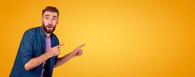 Duygusal beyaz adam turuncu stüdyo arka planında boş bir alanı işaret ediyor, afiş tasarımı. Şok olmuş genç adamın portresi ürün tanıtımı, satışı veya indirimi sunuyor.