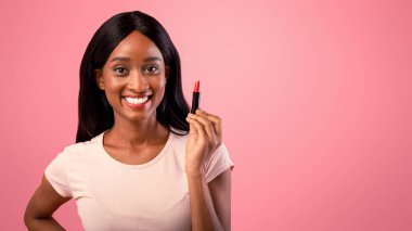 Pembe stüdyo arka planında ruj tutan çekici siyahi kadın. Milenyum Afrikalı Amerikalı kadın elinde makyaj tüpü tutuyor, kozmetik ürünler uyguluyor, dudak parlatıcısını destekliyor.