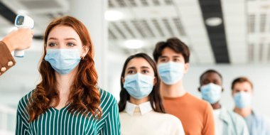 Covid-19 salgını sırasında sabah işte. Temassız lazer silahı termometresi olan kişi modern ofisin girişinde koruyucu maskeli çok ırklı çalışanların vücut ısısını ölçer.
