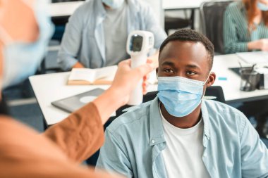 Önlem ve Önlem. Dijital tıbbi temas olmayan kızılötesi termometre tabancası kullanan kadın, cerrahi maske takan Afrikalı Amerikalı bir öğrencinin vücut sıcaklığını ölçüyor ve tarıyor.