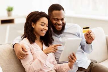 Kolay ödeme. Gülümseyen genç siyah çift bankamatik kartı tutuyor ve dijital tablet kullanıyor, hediyeler, giysiler veya ürünler seçiyor, online ödeme yapıyor, oturma odasındaki kanepede dinleniyor.