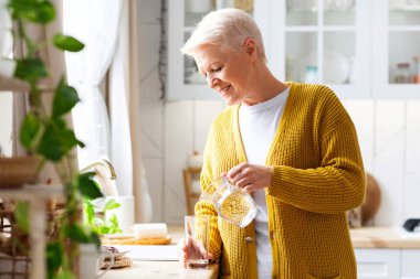 Sıvı kaybı, sağlıklı yaşam tarzı. Neşeli yaşlı bayan bardağa su dolduruyor, mutfak içi. Gayri resmi kıyafetli yakışıklı bir kadın elinde bir bardak tatlı su, kopyalama alanı.