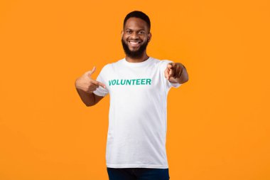 Happy black male volunteer activist pointing finger at his t-shirt and at camera posing on yellow studio background, inviting you to join volunteering career and work. Youre next concept