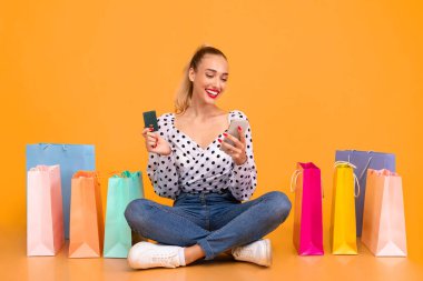 Çevrimiçi Ödeme Kavramı. Elinde kredi kartı olan, gülümseyen şık sarışının portresi, akıllı telefon kullanımı, internetten sipariş vermesi, kıyafet alması, alışveriş torbalarıyla stüdyoda oturması.