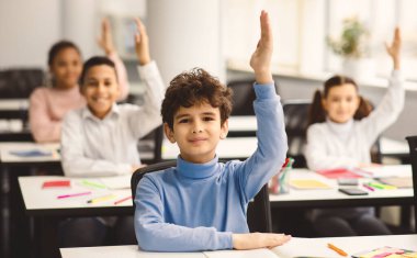 Okulun ilk günü. Sınıfta masada oturan sevimli bir çocuğun portresi, bir grup çok kültürlü küçük öğrenci cevap için ellerini kaldırıyor, modern sınıfta çalışıyor, seçici bir odak noktası var.