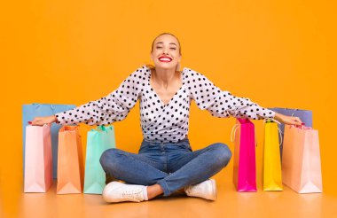 Alışverişe bayılırım. Portakal rengi stüdyo arka plan duvarında izole edilmiş renkli alışveriş torbaları arasında bacak bacak üstüne atmış neşeli, şık bir bayanın portresi.