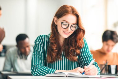 Eğitim, Lise, Öğrenme ve İnsanlar Konsepti. Gözlüklü güzel bir kız öğrencinin portresi. Sınıfta yazı sınavı. Sınıf arkadaşlarıyla aynı masada oturuyor.