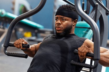Motive olmuş genç Afro-Amerikalı adam spor salonunda göğüs pres makinesi üzerinde çalışıyor, ağırlık kaldırıyor, kollarını ve gövde kaslarını geliştiriyor, yakın plan, fotokopi odasına bakıyor.