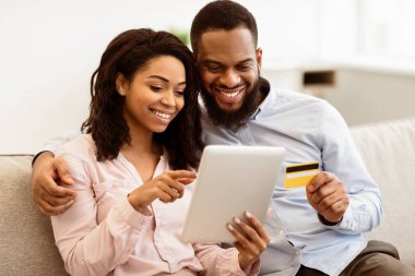 Çevrimiçi Alışveriş ve Ticaret. İki kişilik mutlu Afro-Amerikan ailesi evlerinde kanepede oturuyor, internetten kıyafet veya ürün seçiyor ve dijital tablet kullanarak kredi kartıyla ödeme yapıyorlar.