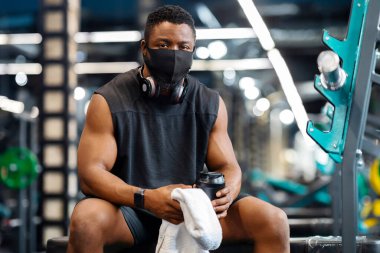 Salgın sırasında spor, önce güvenlik. Spor salonunda egzersiz yaparken koruyucu maske takan, protein içeceği ve havlusu tutan kablosuz kulaklık takan Afrika kökenli Amerikalı sporcu.