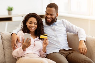 Kolay Ödeme Konsepti. Mutlu Afro-Amerikan erkek ve kadın portresi cep telefonu kullanıyor, kredi kartı tutuyor, evdeki kanepede oturuyor, kocasını kucaklıyor. Perakende ve Satın Al