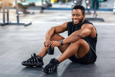 Spor giyim bölümünde gülümseyen siyah bir adam spor salonunda yerde oturmuş protein içeceği içiyor ve mekanı kopyalıyor. Neşeli Afro-Amerikan sporcusu çalışma sırasında mola veriyor, elinde su şişesi tutuyor.