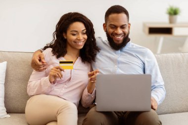 Çevrimiçi Aile Alışveriş. Gülümseyen siyah çift dizüstü bilgisayar kullanıyor ve internetten alışveriş yapıyor, hediyeler sipariş ediyor, kredi kartıyla ödeme yapıyor, oturma odasında kanepede oturuyor.