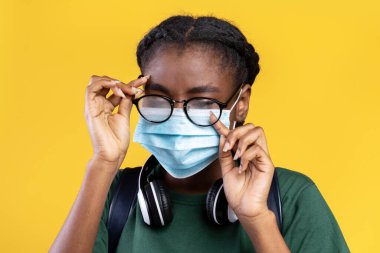 Dont Touch Your Face. Black Female Student Wearing Medical Mask And Scratching Eye Standing Over Yellow Studio Background. Covid-19 Pandemic Outbreak, Coronavirus Protection