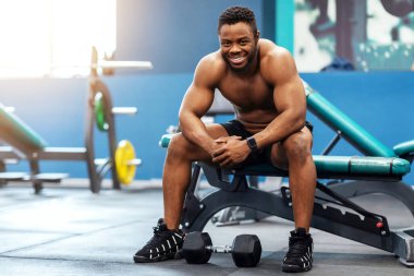 Neşeli tişörtsüz siyah adam bankta oturuyor ve kameraya gülümsüyor, pazılar çalıştıktan sonra dinleniyor, jimnastik salonu içi, fotokopi odası. Mutlu Afro-Amerikan sporcusu halterli egzersizlerin keyfini çıkarıyor.