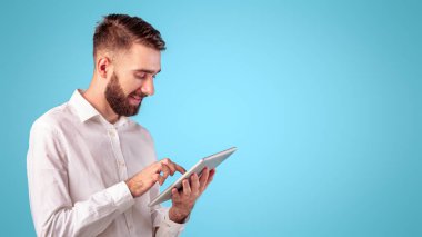 Ofisteki kendine güvenen genç adam mavi stüdyo geçmişi yerine tablet bilgisayar kullanıyor. Odaklanmış serbest çalışan ya da uzaktan çalışan, web konferansı yapan, internetten iletişim kuran