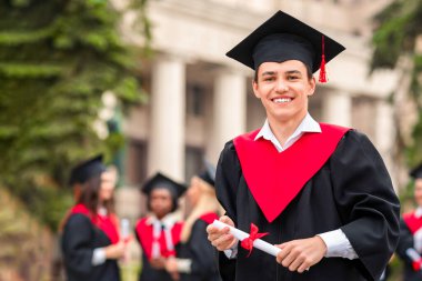 Siyahi ve kırmızı mezuniyet kostümü giymiş yakışıklı bir öğrenci. Diplomasını giymiş ve kameraya gülümsüyor. Üniversite kampüsünde sohbet eden uluslararası öğrencilerin başında dikiliyor.