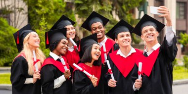 Mezuniyet elbisesi giymiş, akıllı telefondan selfie çeken, diploma taşıyan ve üniversite binasında poz veren, mezuniyeti kutlayan çok ırklı bir grup pozitif öğrenci.