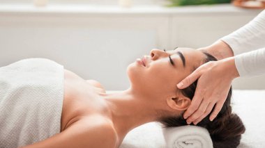 Rahatlamış Asyalı kadın spa salonunda kafa masajı yaptırıyor, güzellik terapisinden zevk alıyor, kapalı gözlerle masaya uzanıyor, kozmetoloji dolabında sağlık gününün tadını çıkarıyor, boş alanla yakın çekim yapıyor.