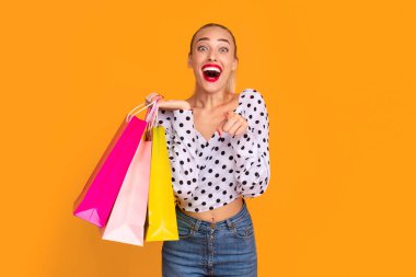 Heyecanlı, modaya uygun, ağzı açık, kamerayı sana çeviren ve elinde parlak renkli kağıt torbaları olan, turuncu arka plan duvarında izole edilmiş bir stüdyoda duran,