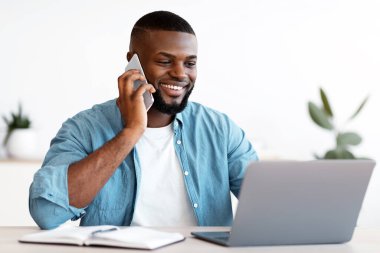 Siyah erkek girişimci cep telefonuyla konuşuyor ve ofiste dizüstü bilgisayarla çalışıyor. Gülümseyen Afrikalı Afrikalı Amerikalı İşadamı bilgisayarla masada oturuyor ve cep telefonu kullanıyor.