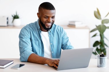 Mesafe işi. Milenyum Afrikalı Amerikalı adam İçişleri Bakanlığı 'nda bilgisayarda çalışıyor. Yakışıklı, gülümseyen, serbest çalışan, masada oturan ve dizüstü bilgisayara bakan adam.