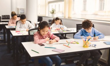 Okul Konsepti. Okulda çeşitli etnik çeşitlilikte çocukların okuması, sosyal mesafeli masalarda oturması, not alması ve kitap yazması. Uluslararası Öğrenciler Yeni Malzeme Öğreniyor