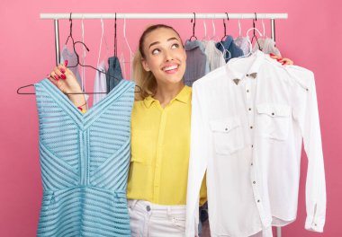 İkilem kavramı. Elbise ve tişörtle askıları tutan, alışverişi düşünen, ne giyeceğine karar veren, arka planda askılık takan genç düşünceli bir kadının portresi.