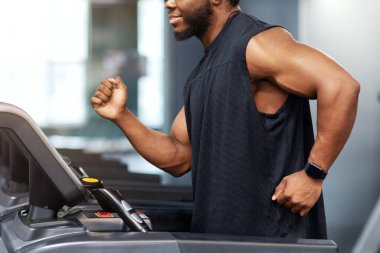 Siyah spor giyim giymiş Afro-Amerikalı bir adam spor salonunda koşuyor, antrenman öncesi ısınıyor. Koşu bandında koşan, boş alana bakan mutlu siyahi adamın yan görüntüsü