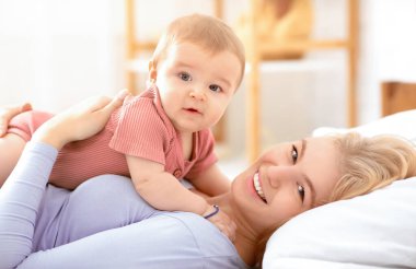 Güzel küçük kız bebek, mutlu sarışın anne göbeğine uzanıyor ve kameraya, yan görüntüye, kopyalama alanına bakıyor. Neşeli bekar anne, küçük tatlı çocuğuyla evde, yatak odasının içinde iyi vakit geçiriyor.