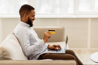 Kolay Ödeme ve Nakit Konsepti. Elinde kredi kartı tutan ve dizüstü bilgisayar kullanan gülümseyen Afro-Amerikan gençlerinin yan görünüm portresi, evdeki koltukta oturup, klavyeye yazı yazması, boşluğu kopyalaması.