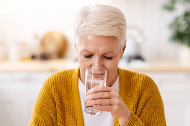 Güzel görünümlü yaşlı bayan mutfakta vakit geçirirken evde su içiyor, fotokopi odası. Beyaz saçlı yaşlı kadın bir bardak su içiyor, sağlıklı kalmak için vücudunu suluyor.