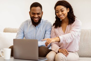 İnternetten alışveriş. Pozitif siyah çift bilgisayar kullanıyor, evde hediyeleri birlikte seçiyorlar. Mutlu siyah kadın dizüstü bilgisayarı işaret ediyor, oturma odasında kanepede ya da evdeki yatak odasında bir adamla oturuyor.