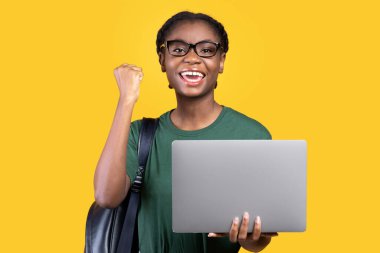 Dizüstü bilgisayarlı neşeli siyah kız öğrenci başarılı çevrimiçi sınavdan sonra sarı stüdyoda poz veriyor. E-Öğrenim, Eğitim Malzemeleri, Uzaktan Çalışma Konsepti