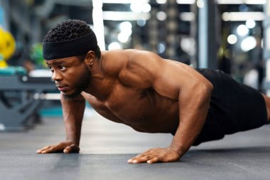 Modern erkek spor salonunda yerde şınav çeken kaslı, üstsüz bir zenci. Motive olmuş Afro-Amerikalı vücut geliştirici. Yerde silah ve göğüs işi yapıyor.