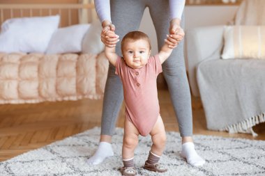 Sevimli küçük bebek ya da kız annesinin yardımıyla ilk adımlarını atıyor, iç mekan, kapat. Anneyle el ele tutuşan sevimli bebek, küçük sarışın kız ya da oğlu oturma odasının önünden geçiyor.