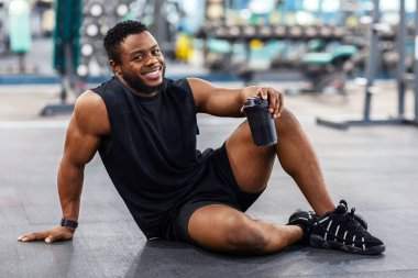 Mutlu Afrikalı Amerikalı vücut geliştirici elinde su ya da spor içeceği bardağıyla spor salonunda yerde dinleniyor ve boş bir alan tutuyor. Gülümseyen siyah kaslı adam modern spor salonunda egzersiz yaparken mola veriyor.