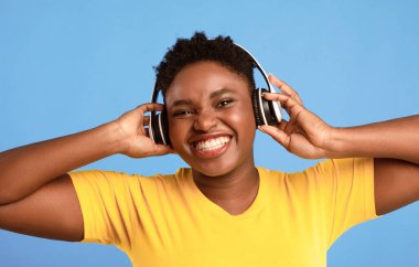 Kablosuz kulaklık takan, Müzik dinleyen, Mavi Stüdyo Arkaplanı Üzerine Gülümseyen Büyük boy Siyah Kadın. Harika Parça Listesi, Elektronik, Müzik Aşığı Konsepti