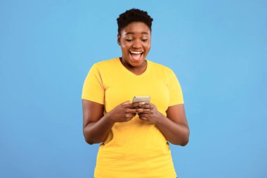 Heyecanlı Afrikalı Büyük Boy Kadın Telefon Uygulaması Mavi Stüdyo Arkaplanının Üzerinde Duruyor. Siyah kadın cep telefonuyla cep telefonu oyunu oynuyor. Akıllı telefon kavramın için harika bir uygulama.