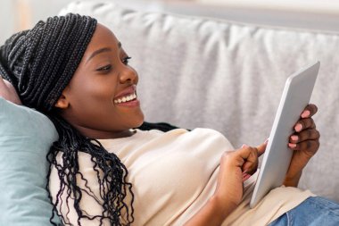 Neşeli siyahi kadın film izliyor, cep telefonu uygulaması kullanıyor. Rahat Afro-Amerikan genç bayan kanepeye uzanmış dijital tablet kullanıyor ve patlamış mısır yiyor.