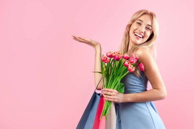 Kadın Günü için özel bir teklif. Güzel elbiseli, elinde çiçekler ve alışveriş çantaları olan güzel sarışın kadın pembe stüdyo arka planında ürününün reklamını yapıyor, tasarım için model yapıyor.