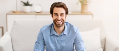 Kanepede oturan ve evdeki kameraya gülümseyen yakışıklı adam. Başarılı Millennial Serbest Çalışan Adam Salonda Poz Veriyor. Kendinden emin Erkek Portresi Konsepti