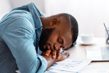 Yorgun Afrikalı Amerikalı serbest çalışan bir adam dizüstü bilgisayarla ve kağıtlarla uyuyor, genç siyahi bir adam iş yerinde uyuyor, yorgun bir erkek elleri üzerinde, uzun bir çalışmadan sonra yorgun düşmüş.