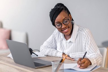 Resmi kıyafetli ve gözlüklü neşeli Afro-Amerikan iş kadını Webinar ya da iş eğitimine katılıyor, çalışma masasında oturuyor, dizüstü bilgisayarını evde kullanıyor, notlar alıyor, yer kopyalıyor.