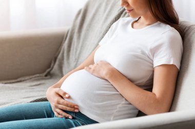 Doğum öncesi zamanı. Güzel müstakbel anne koltukta oturup karnını kucaklıyor, mutlu hamile kadın oturma odasında koltukta dinleniyor, şefkatle karnına dokunuyor ve gülümsüyor, yan görünüm, boş alan.