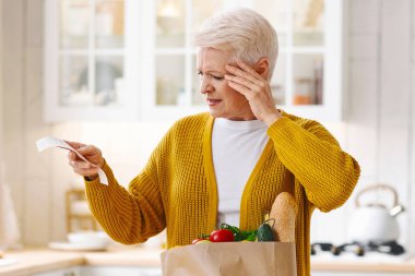 Yaşlı bir kadın, elinde poşetle market faturasını kontrol edip kafasına dokunmuş, mutfakta tek başına durmuş, fotokopi çekiyordu. Yaşlı kadın marketin yüksek fiyatlarından rahatsız oldu.