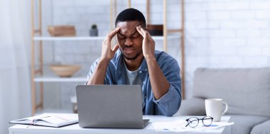 Afro-Amerikalı iş adamı dizüstü bilgisayarında evde çalışırken baş ağrısı çekiyor. Kapalı alanda migren ağrısı çeken Siyah Adam Şakaklarına Masaj Yapıyor. Seçici Odaklanma