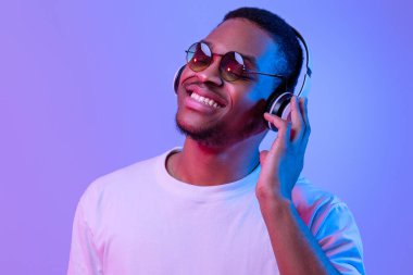 Kablosuz kulaklık takan ve güneş gözlüğü takan olumlu siyahi adam, Mor Arkaplan 'ın altında dururken en sevdiği şarkıları dinleyen Afrikalı genç adam,